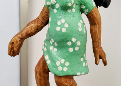 Holzfigur einer lächelnden Frau mit dunklen Haaren, einem grünen Kleid mit weißen Punkten und roten High Heels. Die Figur steht auf einem Sockel und wirkt, als sei sie in Bewegung. Sägearbeit der Künstlerin Ragna Reusch.