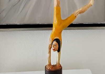 Holzskulptur einer Frau im Handstand auf einem runden Sockel. Sie trägt einen gelben Ganzkörperanzug, ihr schwarzes Haar fällt nach unten. Die Beine sind dynamisch gespreizt, das Gesicht zeigt ein Lächeln. Sägearbeit der Künstlerin Ragna Reusch.