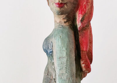Holzskulptur einer Frau mit langen roten Haaren, die über eine Schulter nach vorn fallen. Die Figur ist seitlich dargestellt, mit leichtem Lächeln und Blick zur Seite. Der Körper ist in natürlichen Farbtönen gehalten, mit angedeuteter Bekleidung. Sägearbeit der Künstlerin Ragna Reusch.