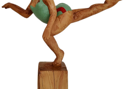 Holzfigur einer Frau in sportlicher Pose mit ausgestrecktem Bein nach hinten, balancierend auf einem Bein auf einem Holzsockel. Sie trägt einen türkisfarbenen Badeanzug mit roten Streifen und hat dunkle, zurückgekämmte Haare. Die Haltung erinnert an eine Turn- oder Ballettübung. Sägearbeit der Künstlerin Ragna Reusch.