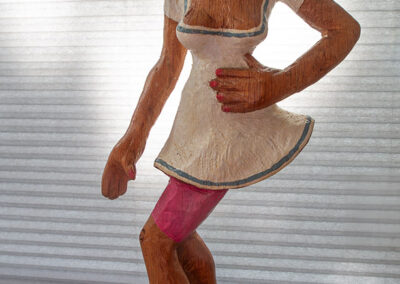Holzskulptur einer Frau mit kurzem blondem Haar, die ein kurzes weißes Kleid mit tiefem Ausschnitt und pinke Strumpfhosen trägt. Sie steht auf roten High Heels und wirkt in einer tänzerischen Bewegung. Die Figur hat eine Hand an der Taille, die andere locker seitlich. Sägearbeit der Künstlerin Ragna Reusch.